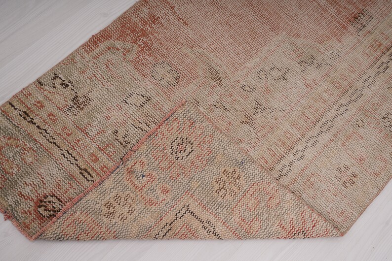May include: A close-up of a folded, woven rug with a beige and pink color scheme. The rug has a geometric pattern with black accents.