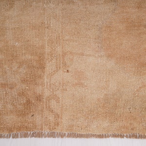 May include: A close-up of a light brown woven rug with a fringe border. The rug has a subtle texture and a slightly faded appearance.