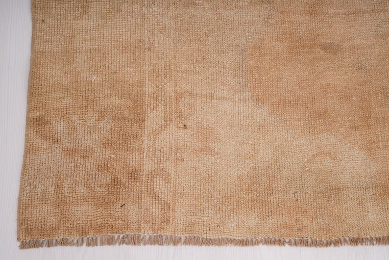 May include: A close-up of a light brown woven rug with a fringe border. The rug has a subtle texture and a slightly faded appearance.