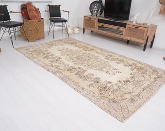 Op de afbeelding: Een rechthoekig, beige en bruin patroonkleed met bloemen- en krulontwerpen. Het kleed ligt op een witte vloer en het patroon is over het gehele oppervlak zichtbaar. Het kleed is een blikvanger in een kamer met meubels.
