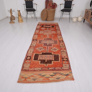 May include: An orange and brown vintage runner rug with geometric patterns. The rug features a central design with dark brown and tan shapes, surrounded by a lighter orange field. The edges have a patterned border with triangles and other shapes.