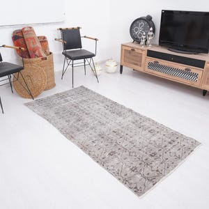 Puede incluir: Una alfombra rectangular gris claro con un estampado floral está colocada sobre un suelo blanco. La alfombra es el punto focal de la habitación, con un aspecto desgastado. Otros muebles incluyen sillas negras, un mueble de TV de madera y una estatua de caballo dorada.