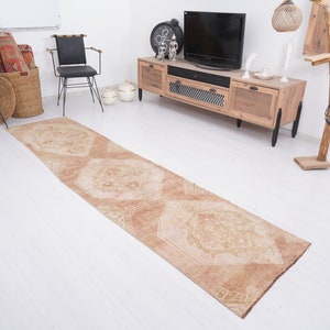 May include: A light brown runner rug with a geometric pattern. The rug is laid out on a white wood floor in a living room setting.