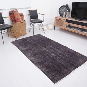 Puede incluir: Una alfombra rectangular gris oscuro con un aspecto envejecido y vintage. La alfombra tiene un patr&oacute;n descolorido y moteado con reflejos grises m&aacute;s claros. La alfombra est&aacute; colocada sobre un suelo blanco, y la habitaci&oacute;n incluye muebles y objetos decorativos.