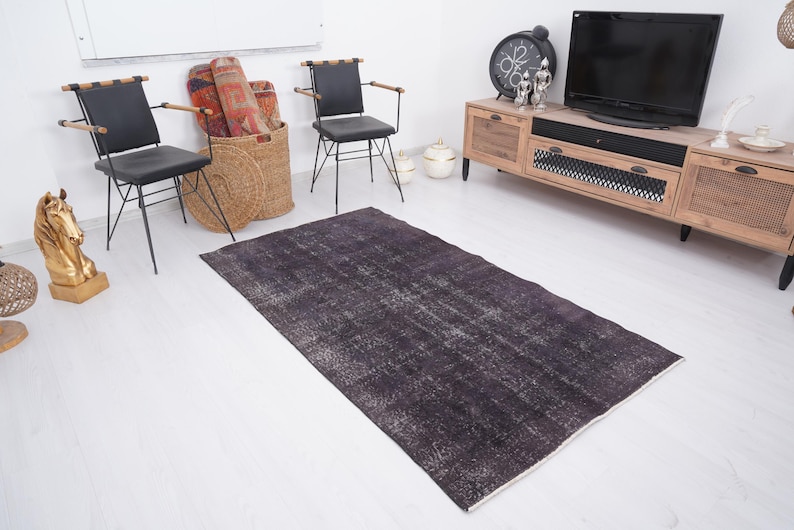 Puede incluir: Una alfombra rectangular gris oscuro con un aspecto envejecido y vintage. La alfombra tiene un patr&oacute;n descolorido y moteado con reflejos grises m&aacute;s claros. La alfombra est&aacute; colocada sobre un suelo blanco, y la habitaci&oacute;n incluye muebles y objetos decorativos.