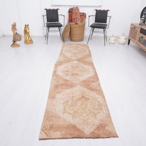 May include: A long, rectangular, beige and brown patterned rug with a geometric design. The rug is laid out on a white floor.