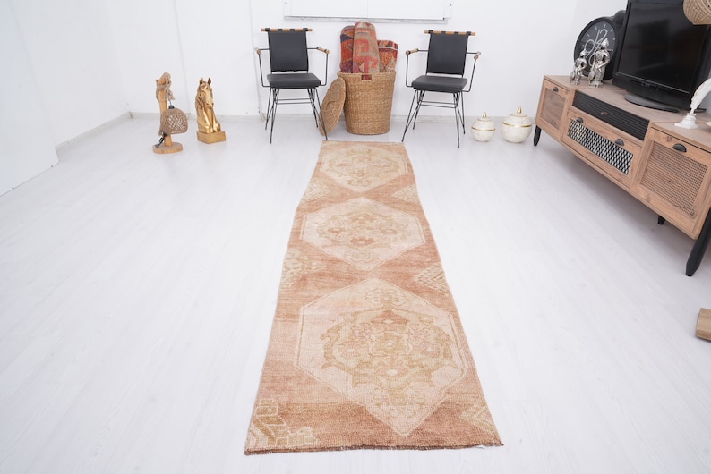May include: A long, rectangular, beige and brown patterned rug with a geometric design. The rug is laid out on a white floor.