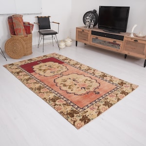 Puede incluir: Una alfombra rectangular con un estampado floral, con un campo central rojo y un diseño de medallón floral beige. La alfombra tiene un borde marrón con detalles florales. Está sobre un suelo blanco.