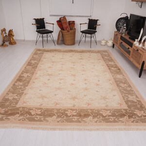 May include: A beige area rug with a floral pattern and a brown border. The rug has a fringe edge and is laid out on a white floor.
