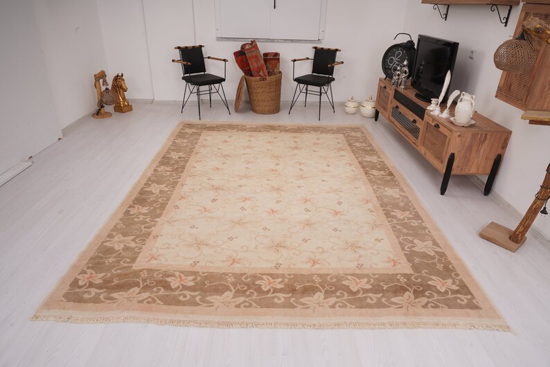 May include: A beige area rug with a floral pattern and a brown border. The rug has a fringe edge and is laid out on a white floor.