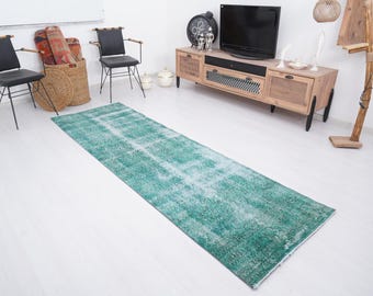 May include: A long, rectangular, distressed green rug with a faded white pattern. The rug is placed on a white floor, with furniture and decor in the background, including chairs, a TV, and a wooden cabinet.