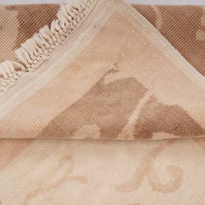 May include: A close-up of a beige and brown rug with a fringe border. The rug has a subtle pattern of geometric shapes.