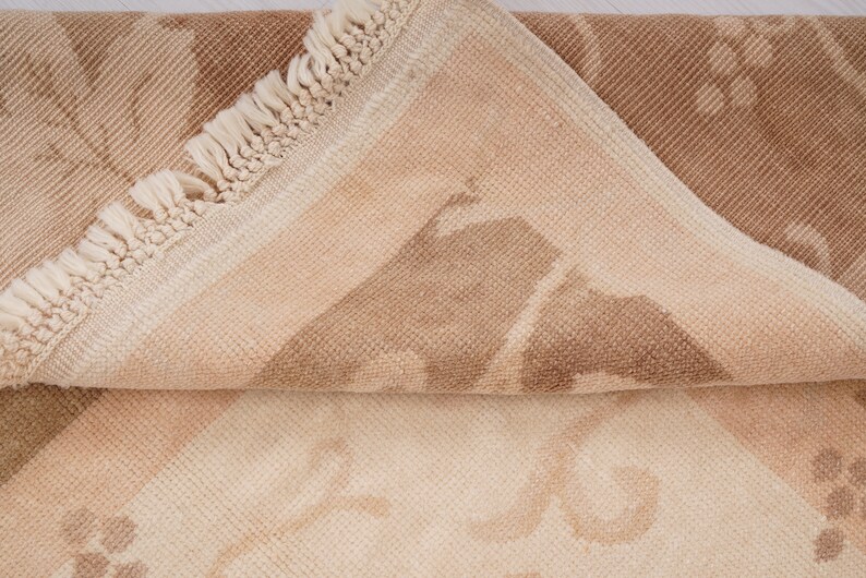 May include: A close-up of a beige and brown rug with a fringe border. The rug has a subtle pattern of geometric shapes.