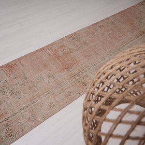 May include: A long, rectangular rug with a faded pink and cream color scheme. The rug has a traditional Persian design with intricate floral patterns. The rug is lying on a white surface.