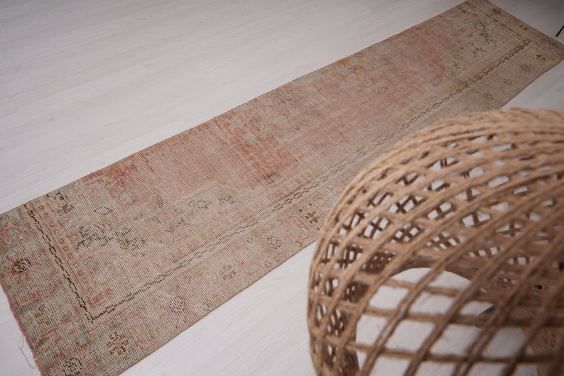 May include: A long, rectangular rug with a faded pink and cream color scheme. The rug has a traditional Persian design with intricate floral patterns. The rug is lying on a white surface.