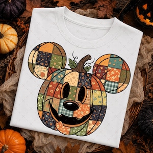 May include: A white t-shirt featuring a large graphic of a pumpkin shaped like a mouse head. The pumpkin is composed of various patchwork fabric patterns in shades of orange, green, and brown, with a carved jack-o'-lantern face and green leaves.