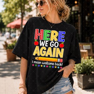 Könnte beinhalten: Ein schwarzes Kurzarm-T-Shirt mit dem bunten Schriftzug HERE WE GO AGAIN, zwei roten Äpfeln, einer gelben Bleistiftgrafik und dem Text I mean welcome back! darunter, gefolgt von einer Reihe kleiner bunter Sterne am unteren Rand.