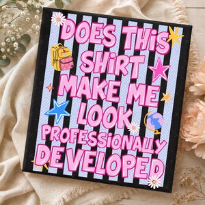 May include: A rectangular sign with a black and light blue striped background featuring the pink text "DOES THIS SHIRT MAKE ME LOOK PROFESSIONALLY DEVELOPED". The design includes illustrations of a yellow backpack, stars, a globe, and daisies.