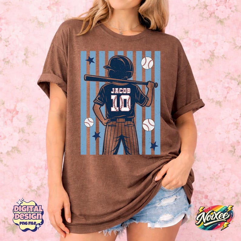 May include: Brown t-shirt featuring a baseball player graphic with the name "Jacob" and the number "10" on the back. The design includes baseballs, stars, and blue stripes. The shirt is a casual style.
