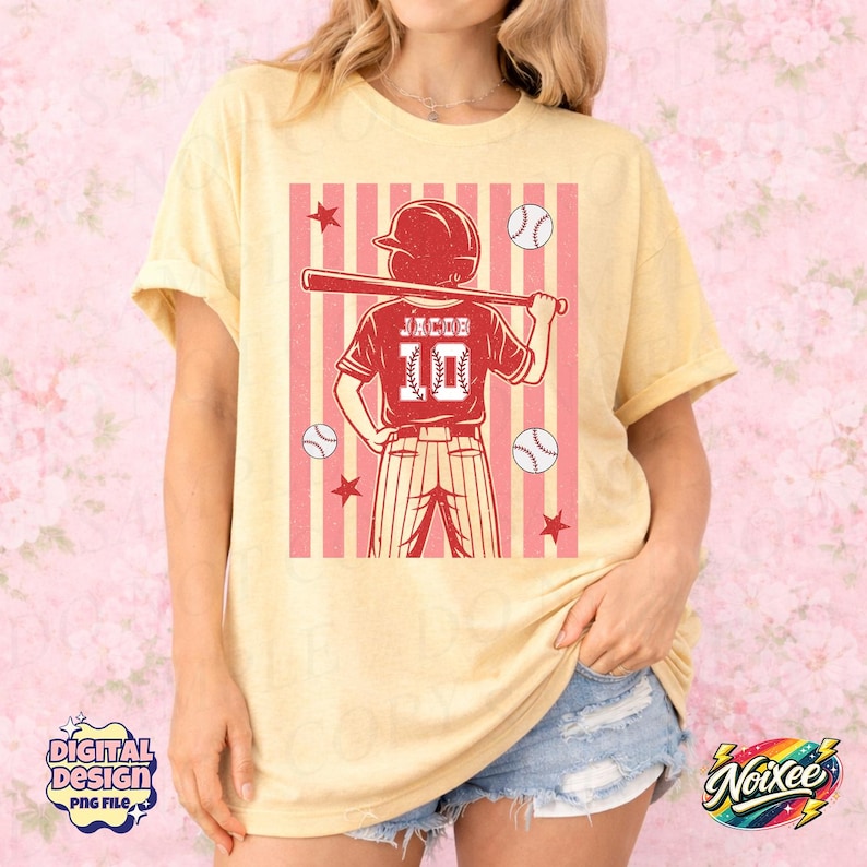 May include: A light yellow t-shirt featuring a baseball-themed graphic. The graphic includes a baseball player holding a bat, baseballs, stars, and the name "Jacob" with the number "10". The design is set against a pink and white striped background.