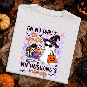 May include: A white t-shirt featuring a graphic of a ghost wearing a witch hat and sunglasses, pushing a shopping cart filled with pumpkins and a skull. The ghost holds an orange jack-o'-lantern bag. Text reads: ON MY WAY to spend MY HUSBAND'S money.