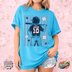 May include: A light blue t-shirt featuring a baseball-themed graphic. The graphic includes a baseball player with a bat, baseballs, stars, and the name "Jacob" with the number "10" on the jersey. The shirt has short sleeves and a relaxed fit.