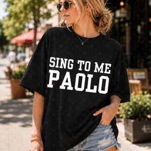May include: A black short-sleeved t-shirt featuring the white text "SING TO ME PAOLO" in a bold, block-style font across the chest. The garment has a crew neckline and a relaxed, loose-fitting silhouette, constructed from a solid black fabric.
