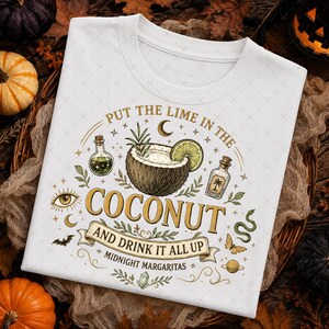 May include: A white t-shirt featuring a graphic print of a coconut, a lime slice, and two glass bottles. The text reads, "PUT THE LIME IN THE COCONUT AND DRINK IT ALL UP" and "MIDNIGHT MARGARITAS," accompanied by illustrations of a crescent moon, eye, bat, snake, butterfly, and planet.