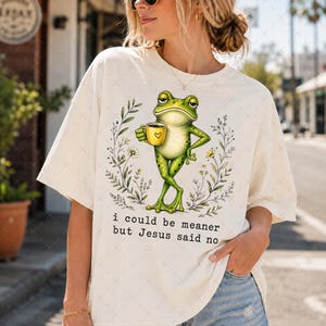 Könnte beinhalten: Ein cremefarbenes T-Shirt mit einer Grafik eines grünen Frosches, der eine gelbe Tasse mit einem Herzsymbol hält. Der Frosch ist von einem Blumenkranz umgeben und der Text "i could be meaner but Jesus said no" steht in schwarzer Schrift darunter.