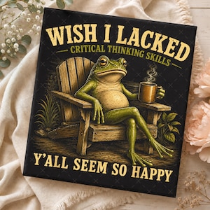Puede incluir: Un cuadro decorativo rectangular de fondo negro con una rana verde sentada en una silla de madera, sosteniendo una taza marrón con vapor. El texto dice WISH I LACKED CRITICAL THINKING SKILLS Y'ALL SEEM SO HAPPY en letras amarillas y negritas.