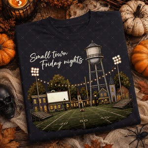 May include: A black crewneck sweatshirt featuring a graphic of a football field at night with a water tower, stadium lights, and the text Small town Friday nights in white script. The scene includes bleachers and a scoreboard.