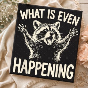 May include: A black square sign featuring a white line art illustration of a raccoon with its paws raised and mouth open in a surprised expression. The text "WHAT IS EVEN HAPPENING" is printed in bold white capital letters above and below the raccoon.