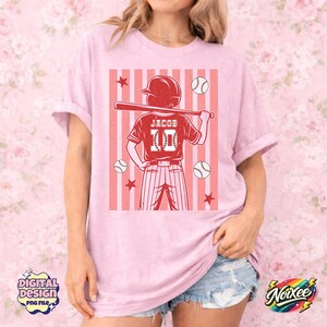 May include: Pink t-shirt featuring a baseball-themed graphic. The graphic includes a baseball player holding a bat, baseballs, stars, and the name "Jacob" with the number "10". The shirt has short sleeves and a relaxed fit.