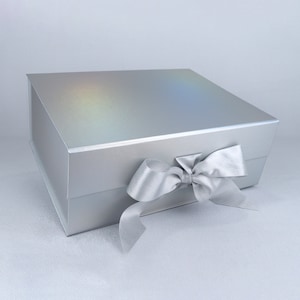May include: A silver gift box with a matching satin ribbon tied in a bow. The box has a magnetic closure and a subtle iridescent sheen on the lid, creating a luxurious appearance. The box is empty.