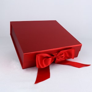 May include: A red gift box with a matching satin ribbon tied in a bow. The box is square-shaped with a glossy finish. The ribbon hangs down on the right side of the box.
