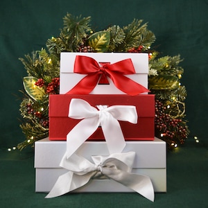 May include: Three gift boxes stacked on top of each other. The top box is white with a red bow. The middle box is red with a white bow. The bottom box is white with a white and silver bow. The boxes are in front of a green background with a Christmas wreath.