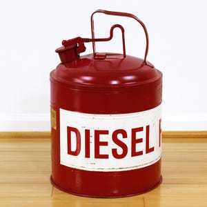 May include: A red metal diesel fuel can with a white band featuring the word "DIESEL" in bold red letters. The can has a handle and a spout with a cap. The can is sitting on a wooden floor.