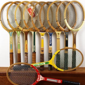May include: A collection of vintage wooden tennis rackets featuring various brands including Spalding, Wilson, and Slazenger. The rackets display diverse color schemes, including red, yellow, and natural wood tones, with branded text and logos.