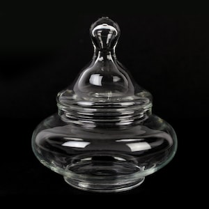 May include: A clear glass jar with a lid. The jar has a rounded base and a bulbous lid with a small handle. The glass is transparent, allowing a view of the contents. The jar is likely used for storage or display.