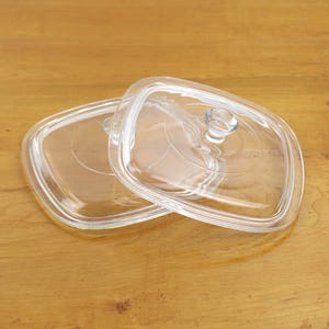 May include: Two clear, square glass lids with rounded corners and small, round handles. The lids are slightly overlapping, and the background is a light brown wooden surface. The lids appear to be for cookware.