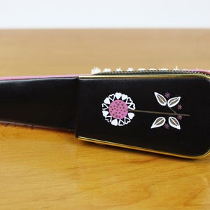 May include: A black, rectangular case with a gold trim and a floral design. The design features a pink and white flower with white leaves. The case is on a wooden surface.