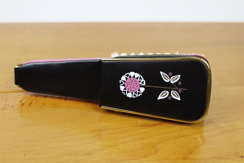 May include: A black, rectangular case with a gold trim and a floral design. The design features a pink and white flower with white leaves. The case is on a wooden surface.