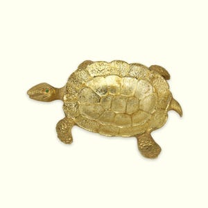 May include: A gold-colored turtle figurine with a textured shell and limbs. The turtle has a small green gem for an eye. The figurine is a decorative item.