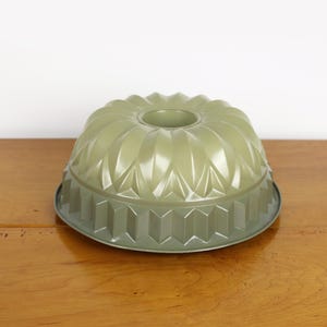 May include: A sage green bundt cake pan with a decorative, fluted design. The metal pan has a circular shape with a central hole and a geometric pattern around the base. The pan sits on a wooden surface.