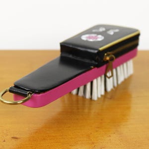 May include: A black and pink brush with a zippered top. The brush has white bristles and a gold-colored zipper and ring. The top of the brush has a small floral design.