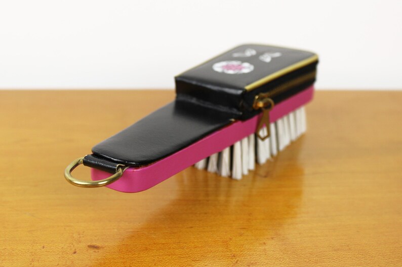 May include: A black and pink brush with a zippered top. The brush has white bristles and a gold-colored zipper and ring. The top of the brush has a small floral design.