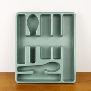 May include: A rectangular, sage green plastic utensil organizer with multiple compartments for silverware. The organizer has slots for forks, knives, and spoons, and is designed to fit inside a kitchen drawer.