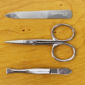 May include: A set of three silver manicure tools: a nail file with the text "Special Triple Cut GERMANY", small scissors, and a cuticle pusher. The tools are arranged on a wooden surface.