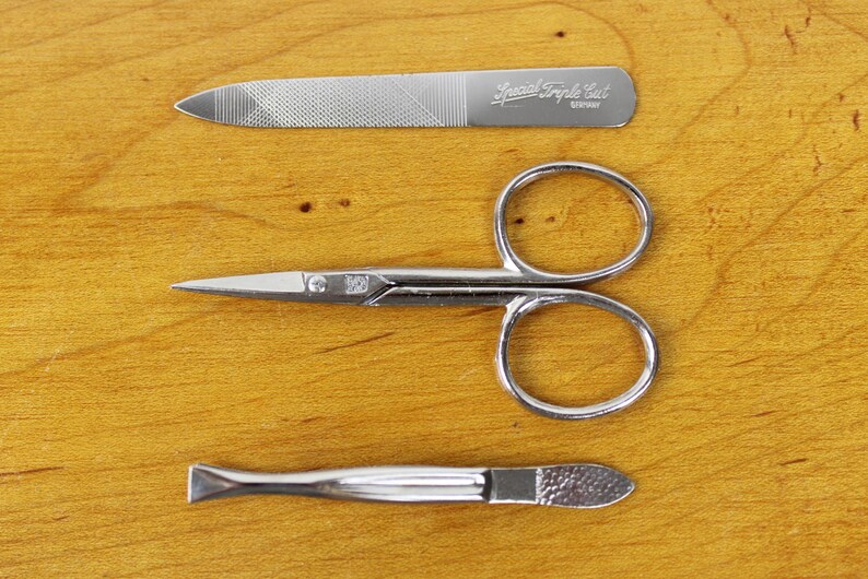 May include: A set of three silver manicure tools: a nail file with the text "Special Triple Cut GERMANY", small scissors, and a cuticle pusher. The tools are arranged on a wooden surface.