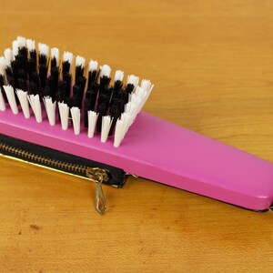 May include: A pink hairbrush with a black zipper case and a gold-colored zipper pull. The brush has black and white bristles and a handle with a gold ring.